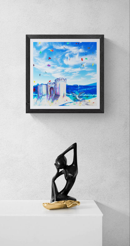 A framed square art print depicting a colorful beach scene with blue sky, seagulls and sandcastle, and confetti displayed on a light gray wall above a white pedestal with a black and gold figurine.