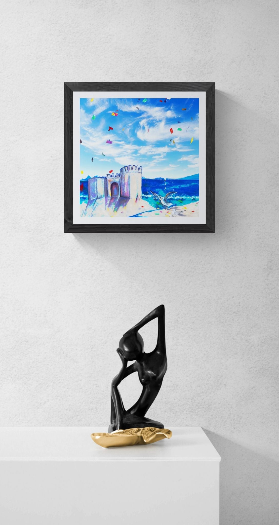 A framed colorful art print of a coastal scene with blue sky, sandcastle, and sea, displayed on a light gray wall above a white pedestal with a black and gold abstract sculpture.