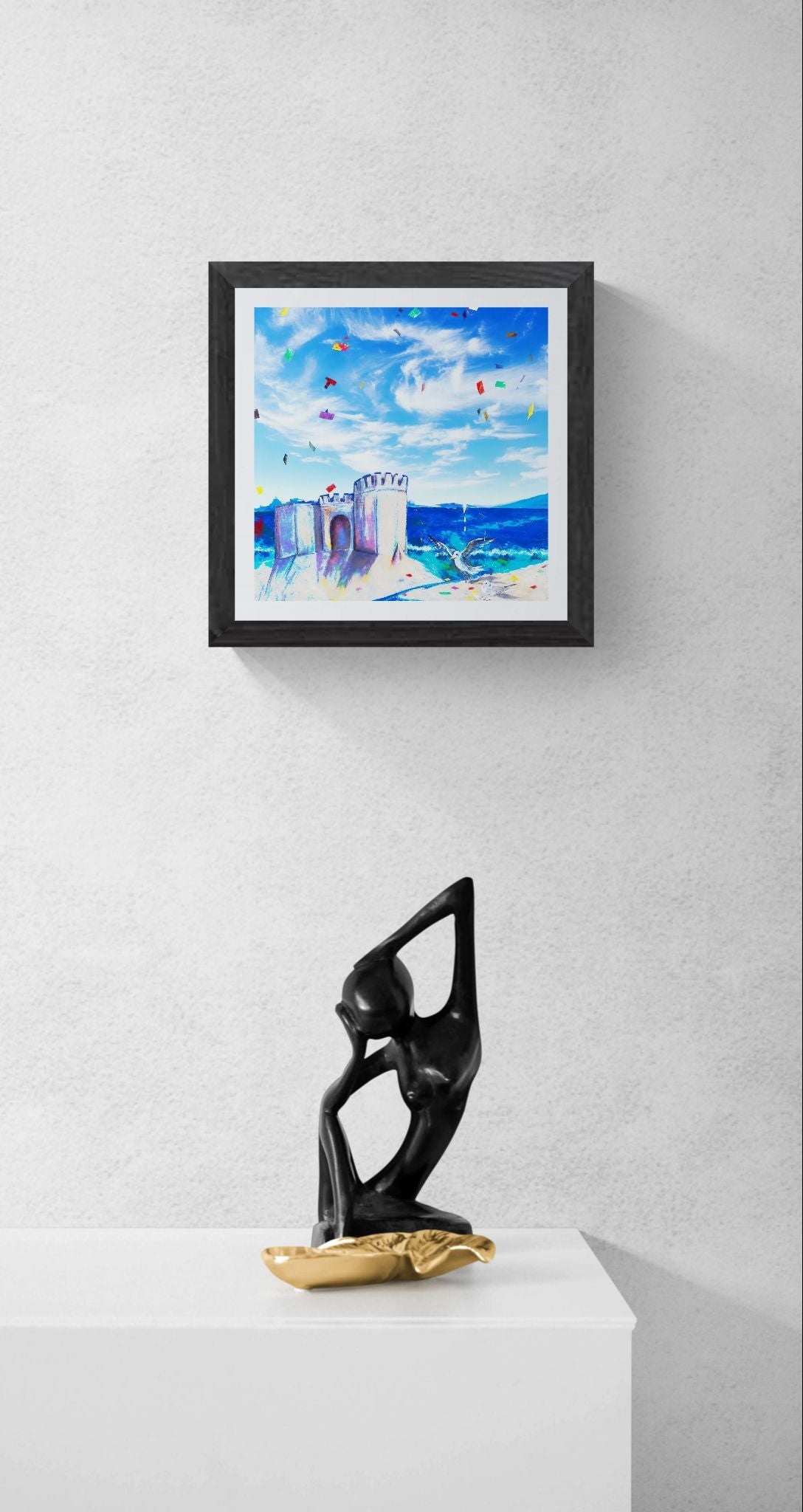 A framed square art print depicting a colorful beach scene with blue sky and a sandcastle, displayed on a light gray wall above a white pedestal with a black and gold abstract sculpture.