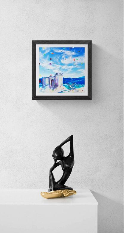 A framed square art print depicting a colorful beach scene with blue sky and a sandcastle, displayed on a light gray wall above a white pedestal with a black and gold abstract sculpture.